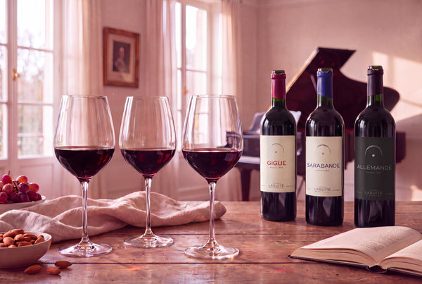 Drei Gläser dunkler Rotwein nebeneinander auf einem Tisch in einem hellen Salon; dahinter die drei Rotweine von LaSuite aux Conseillans — Gigue, Sarabande und Allemande —, im Hintergrund ein geöffneter Flügel.