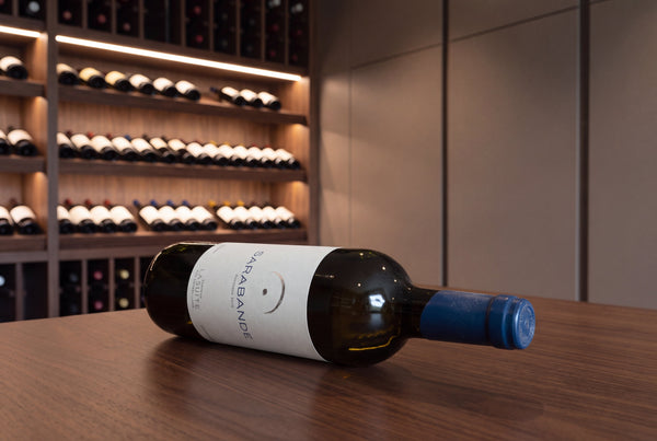 Eine gereifte Flasche Sarabande Bordeaux 2015 liegt auf einem Tisch in einem modernen privaten Weinkeller; dahinter, unscharf, Regale voller Flaschen. Das Etikett ist lesbar.